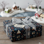 Nautical Christmas gift wrap with sailboats, anchors, and trees, tied with a silver bow and "Seasons Greetings" tag.