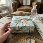 Square gift wrapped in sailboat-patterned paper with twine and tag on a rustic wooden table.