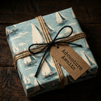 Square gift wrapped in sailboat-patterned paper with twine, black string bow, and "Adventure Awaits" tag.
