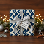 Modern navy blue and cream wave pattern wrapping paper with silver ribbon, tag, and festive Christmas decor on a wood table.