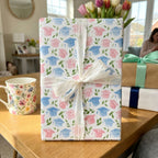 Pink and blue graduation wrapping paper with watercolor caps and white ribbon by Wrapped Studios.