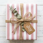 Pink striped wrapping paper with a rustic twine bow and Joy tag on white wood by Wrapped Studios.