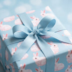 Pink bunny wrapping paper on a baby shower gift box with a blue ribbon bow by Wrapped Studios.