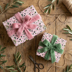 Pink floral wrapping paper with plush green and pink bows on rustic wood. Luxury gift wrap by Wrapped Studios.