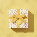 Square gift in floral wrapping paper with yellow ribbon and "Enjoy!" tag. Wrapped Studios.