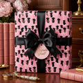 Pink graduation wrapping paper with a black satin bow and "Congrats Grad" tag by Wrapped Studios.