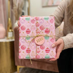 Pink Christmas wrapping paper with cute holiday ornament patterns and a gold bow, from Wrapped Studios.