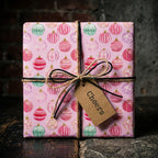 Pink Christmas wrapping paper with holiday ornaments, tied with twine and gift tag by Wrapped Studios.