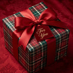 Square gift box wrapped in traditional red and green plaid Christmas gift wrap with red satin bow and tag.