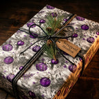 Elegant purple Christmas wrapping paper with ornament pattern, pine sprig, and twine bow by Wrapped Studios.