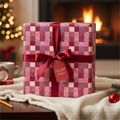 Cozy holiday gift wrapped in red patchwork Christmas wrapping paper with a dark red ribbon and a "Heartfelt Wishes" tag by a warm fireplace.