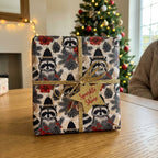 Raccoon Christmas gift wrap with gold glitter ribbon by Wrapped Studios, set against a festive holiday tree.