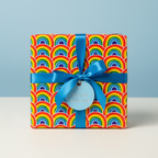 Square gift wrapped in bright rainbow patterned wrapping paper, tied with a blue satin ribbon and tag.