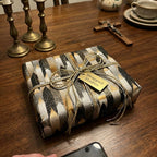Gift wrapped in black, gold, and silver paper on a wooden table with a crucifix and tea set in the background. religious-wrapping-paper-gold-silver-baptism-gift-wrap.