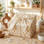 Boho-style gift box wrapped in a wavy checkerboard pattern paper with muted pink and mustard tones, tied with a large cream silk bow.