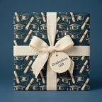 Navy graduation wrapping paper with mortarboard caps and diplomas, tied with a cream bow by Wrapped Studios.