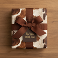 Rustic gift wrapped in cow print paper, tied with brown ribbon and "For You, Wild One" tag, on a wood surface.