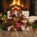 Rustic Christmas gift wrap: Cowhide pattern paper with bronze ribbon and "Happy Trails" tag by cozy fireplace.