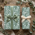 Sage green floral wrapping paper with white wildflowers and elegant bows on rustic wood, by Wrapped Studios.