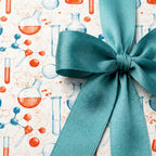 Science wrapping paper with watercolor chemistry beakers and a teal satin bow. By Wrapped Studios.