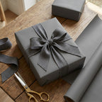 Smoke gray wrapping paper on gift box with a bow on a wooden table with scissors and additional wrapping paper.