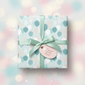 Pastel balloon print wrapping paper on a square gift box, tied with a light green satin bow and a tag.