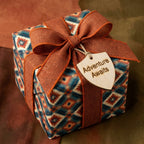 Southwest wrapping paper with a rustic orange bow and 'Adventure Awaits' wood tag by Wrapped Studios.