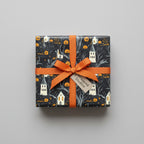 Gift wrapped in black Halloween wrapping paper with spooky houses and pumpkins, tied with an orange ribbon.
