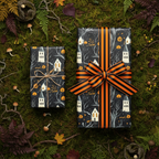 Spooky Halloween gift wrap featuring haunted houses and pumpkins, tied with an orange and black striped ribbon, on a mossy forest floor.