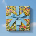 Square gift wrapped in bright yellow floral wrapping paper with light blue ribbon against a blue sky background.