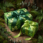 Two presents wrapped in botanical green wrapping paper with ribbon bows on mossy wood.