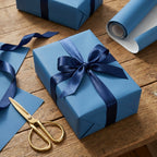 Blue gift box with satin ribbon bow on wood table. Solid color wrapping paper roll and gold scissors.