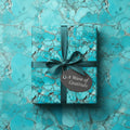 Square gift wrapped in teal stone-patterned paper with ribbon bow and gratitude tag.