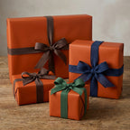 Solid rust-colored gift wrap with brown, navy, and green ribbon bows on wooden table; elegant gift wrapping paper.