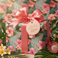 Tropical floral wrapping paper with pink hibiscus, satin bow, and "Aloha Love" tag by Wrapped Studios.