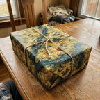 Rectangular gift wrapped in vintage-style toile wrapping paper with blue and yellow tones, tied with rustic twine.