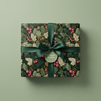 Square gift wrapped in botanical Christmas wrapping paper with dark green ribbon and holly berry design.