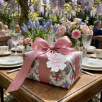 Floral wrapping paper with pink bow and Mother’s Day tag on a garden tea table by Wrapped Studios.