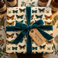 Butterfly wrapping paper on a square gift box, tied with a teal velvet ribbon and vintage-style tag.