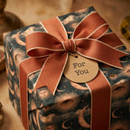 Celestial wrapping paper with vintage moon and sun patterns, tied with a rust bow by Wrapped Studios.