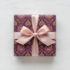 Elegant square gift wrapped in purple floral wrapping paper with light pink ribbon bow on a white wood surface.