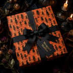 Spooky Halloween wrapping paper with black monsters on orange, tied with a black bow by Wrapped Studios.