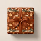 Boho celestial wrapping paper in terracotta with mountains and moons, tied with a rust bow by Wrapped Studios.