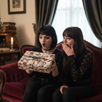 Two women exchange a surprise love-themed gift, wrapped in romantic black and red script paper, on a vintage red velvet couch.