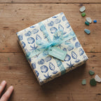 Square gift box wrapped in seashell pattern gift wrap with a light blue ribbon, on a wooden table.