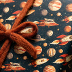 Kids space wrapping paper with retro rockets and planets, tied with an orange yarn bow by Wrapped Studios.