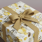 Square gift wrapped in white floral wrapping paper with yellow citrus fruit and gold ribbon.