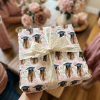 Graduation wrapping paper with watercolor caps, pink flowers, and cream ribbon by Wrapped Studios.