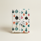 Christmas wrapping paper with watercolor ornaments in teal, black, and orange, tied with a white ribbon and "Wrapped Studios" tag.