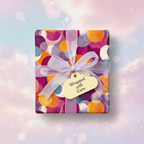Square gift wrapped in colorful geometric wrapping paper with sheer lavender ribbon and a gift tag.